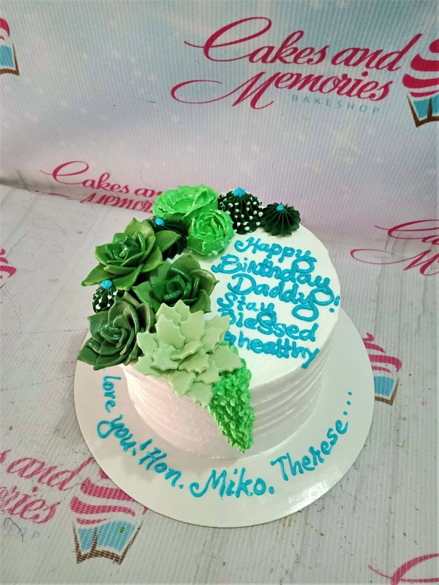 Succulent Garden White Birthday Cake A white and green round 1-tier cake themed as a Succulent Garden, featuring piped icing succulents and cacti, and blue text.