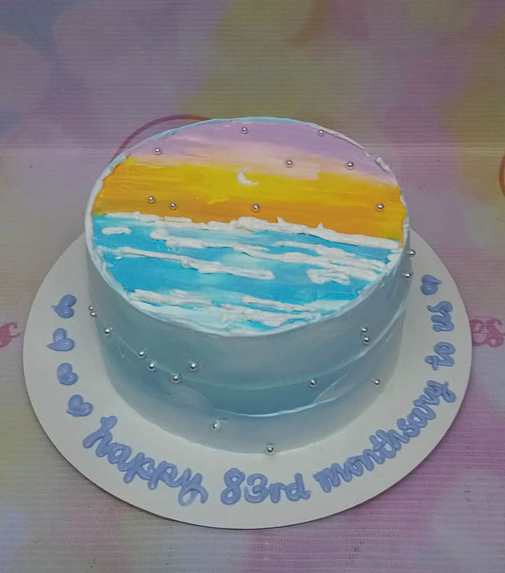 A round minimalist cake featuring a hand-painted sunset beach scene in purple, orange, and blue icing with silver dragees. A round minimalist cake featuring a hand-painted sunset beach scene in purple, orange, and blue icing with silver dragees.
