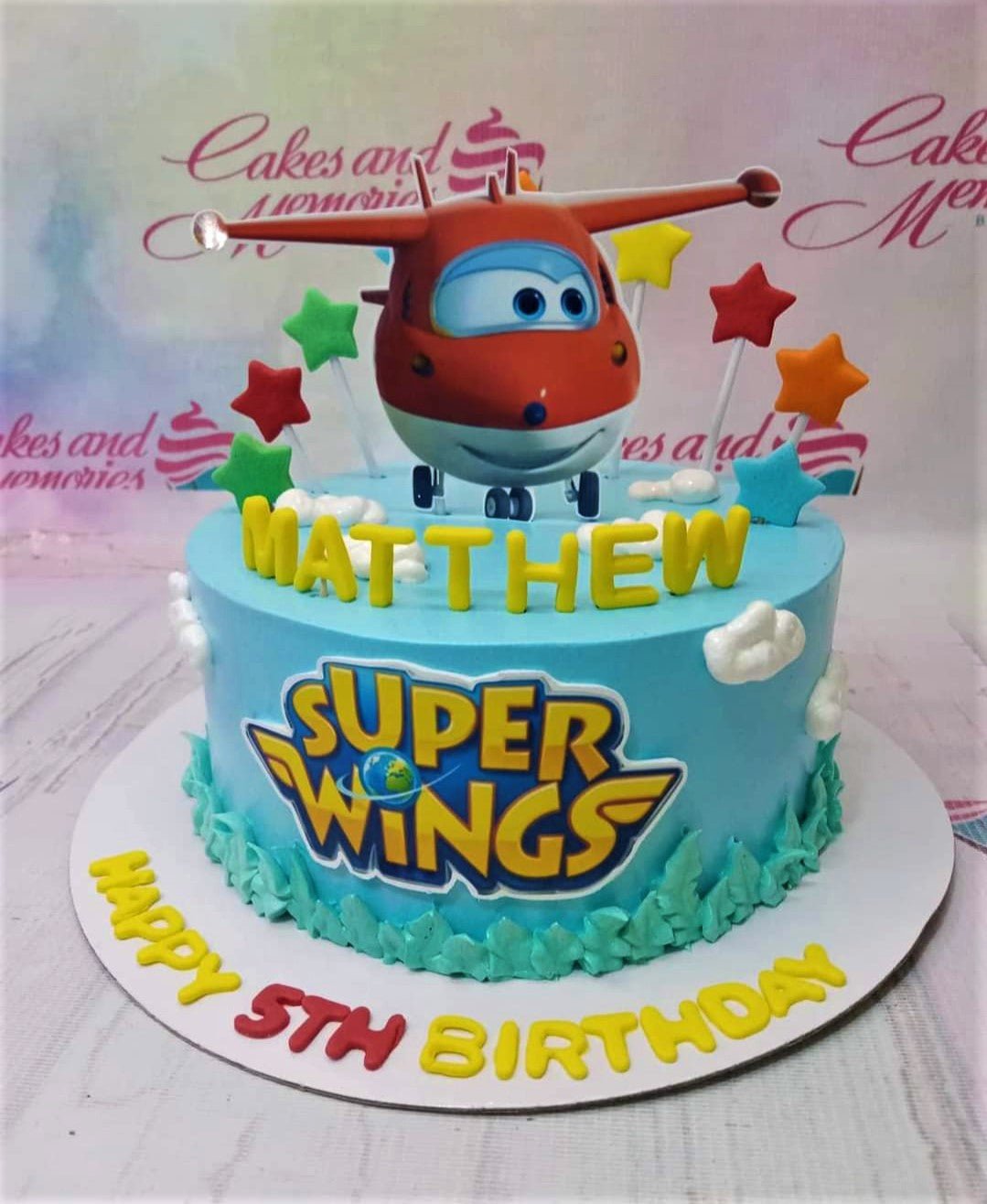 Light blue Super Wings themed birthday cake with a red airplane topper, colorful stars, and yellow gumpaste letters for Matthew's 5th birthday. Light blue Super Wings themed birthday cake with a red airplane topper, colorful stars, and yellow gumpaste letters for Matthew's 5th birthday.