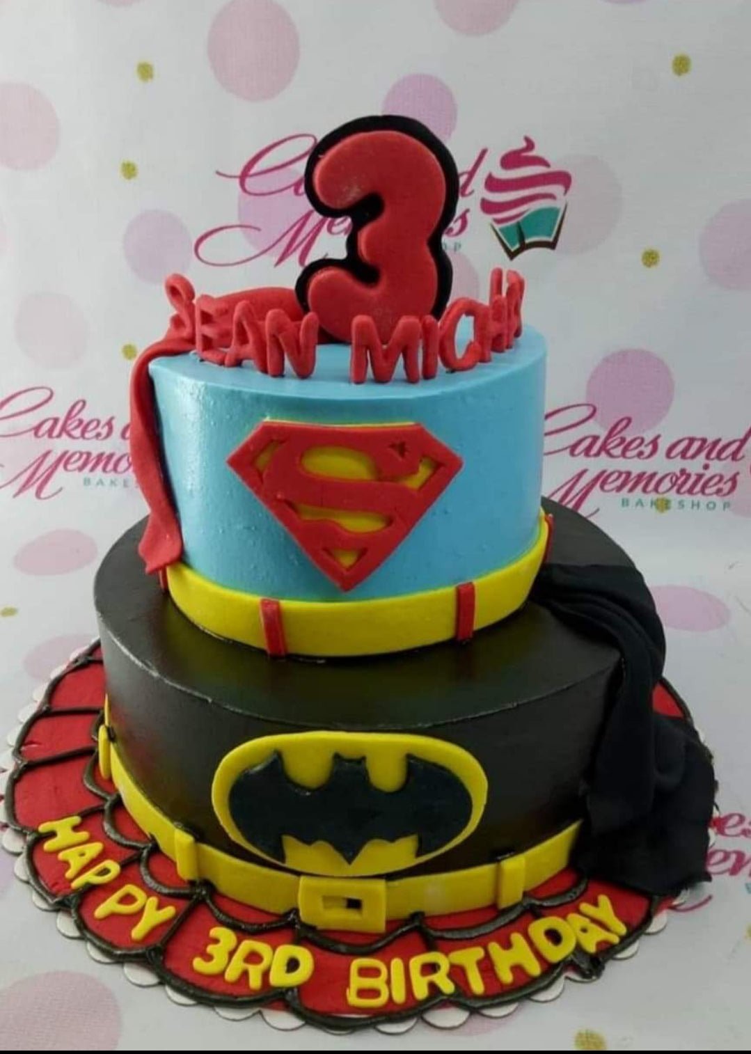 Two-tier superhero themed cake featuring Superman and Batman designs with a red number 3 topper and fondant capes. Two-tier superhero themed cake featuring Superman and Batman designs with a red number 3 topper and fondant capes.