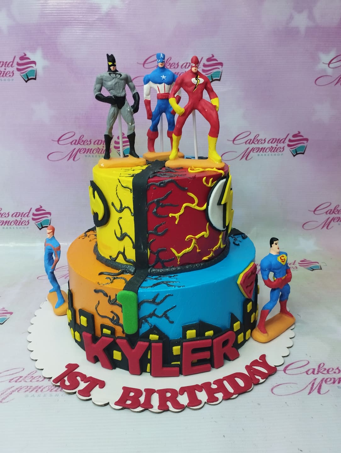 Two-tier superhero themed birthday cake featuring Batman, Captain America, Flash, Superman, and Aquaman figurines with city skyline and comic logos. Two-tier superhero themed birthday cake featuring Batman, Captain America, Flash, Superman, and Aquaman figurines with city skyline and comic logos.