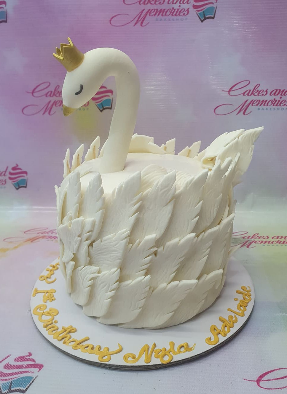 White swan-themed fondant cake with textured gumpaste feathers and a gold crown topper on a white base board with gold lettering.