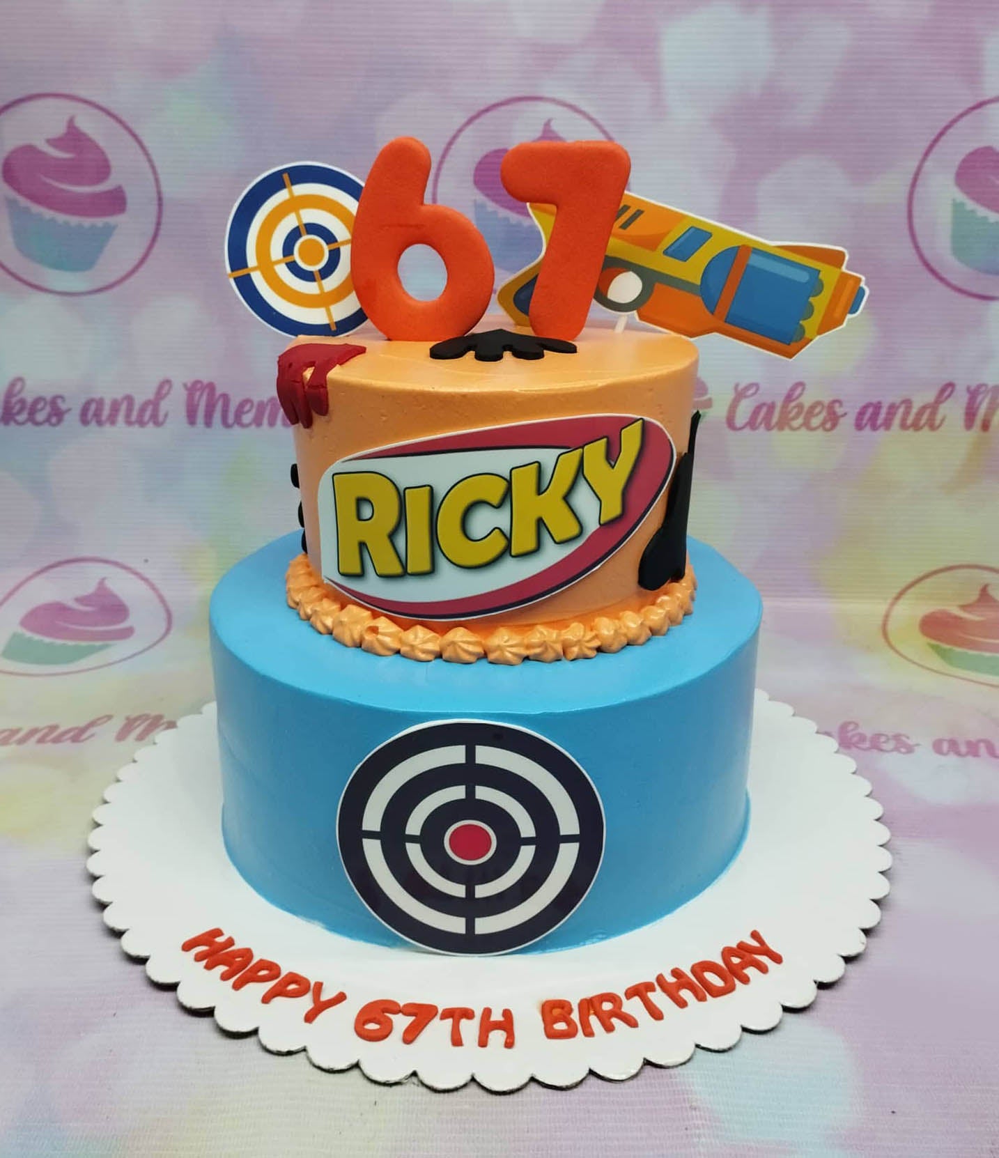 Two-tier orange and blue target themed birthday cake with a toy gun and target printouts for a 67th birthday. Two-tier orange and blue target themed birthday cake with a toy gun and target printouts for a 67th birthday.