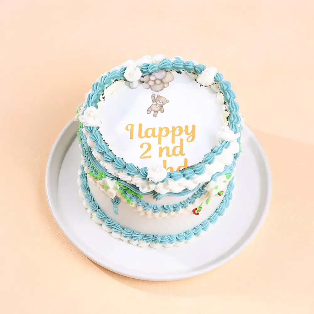 White vintage-style cake with blue and white icing ruffles, featuring a teddy bear printout and Happy 2nd Birthday message. White vintage-style cake with blue and white icing ruffles, featuring a teddy bear printout and Happy 2nd Birthday message.