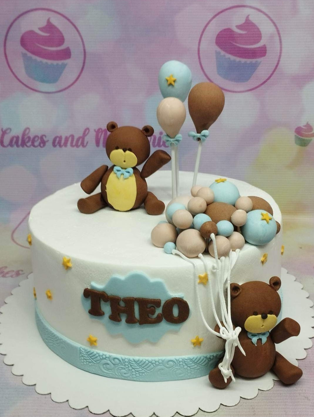 White fondant cake with two brown teddy bears, blue and tan balloons, and a blue name plaque for Theo. White fondant cake with two brown teddy bears, blue and tan balloons, and a blue name plaque for Theo.
