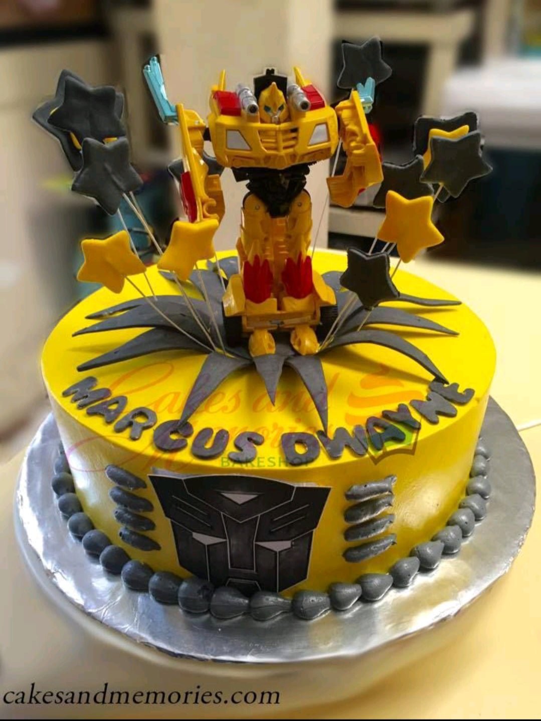 Yellow Transformers Bumblebee themed cake with a 3D robot toy topper, grey fondant stars, and a Transformers logo on the side. Yellow Transformers Bumblebee themed cake with a 3D robot toy topper, grey fondant stars, and a Transformers logo on the side.