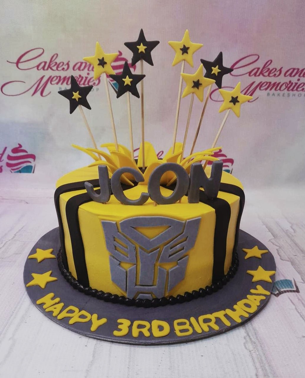 Yellow and black Transformers Bumblebee themed birthday cake with grey Autobot logo and star toppers. Yellow and black Transformers Bumblebee themed birthday cake with grey Autobot logo and star toppers.