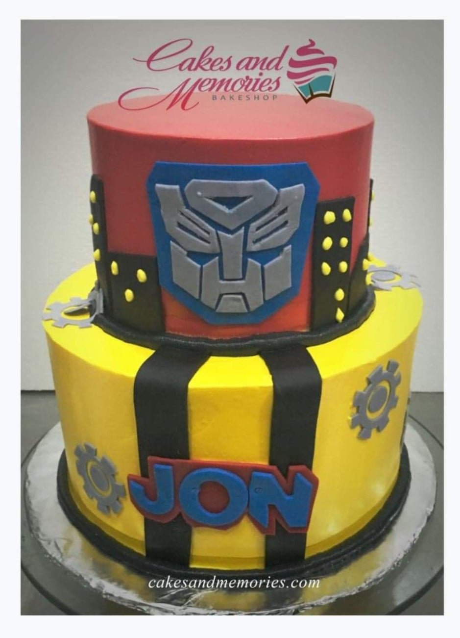 Two-tier Transformers themed birthday cake in red and yellow fondant with Autobot logo and gear decorations. Two-tier Transformers themed birthday cake in red and yellow fondant with Autobot logo and gear decorations.