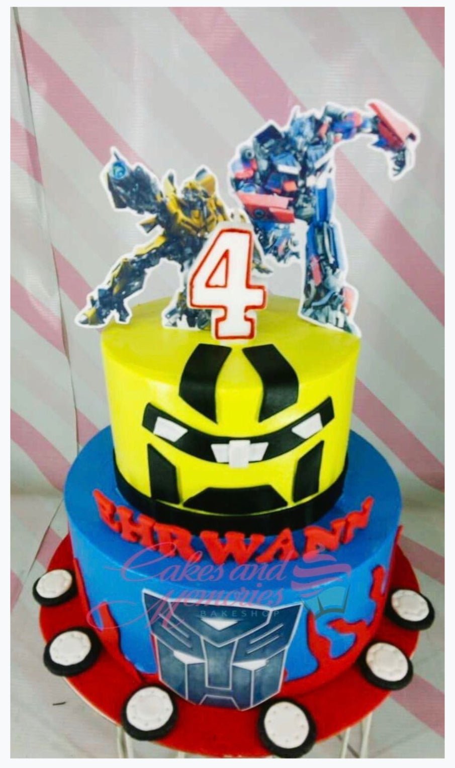 Two-tier Transformers themed birthday cake with yellow Bumblebee top tier and blue Optimus Prime bottom tier featuring character printouts. Two-tier Transformers themed birthday cake with yellow Bumblebee top tier and blue Optimus Prime bottom tier featuring character printouts.