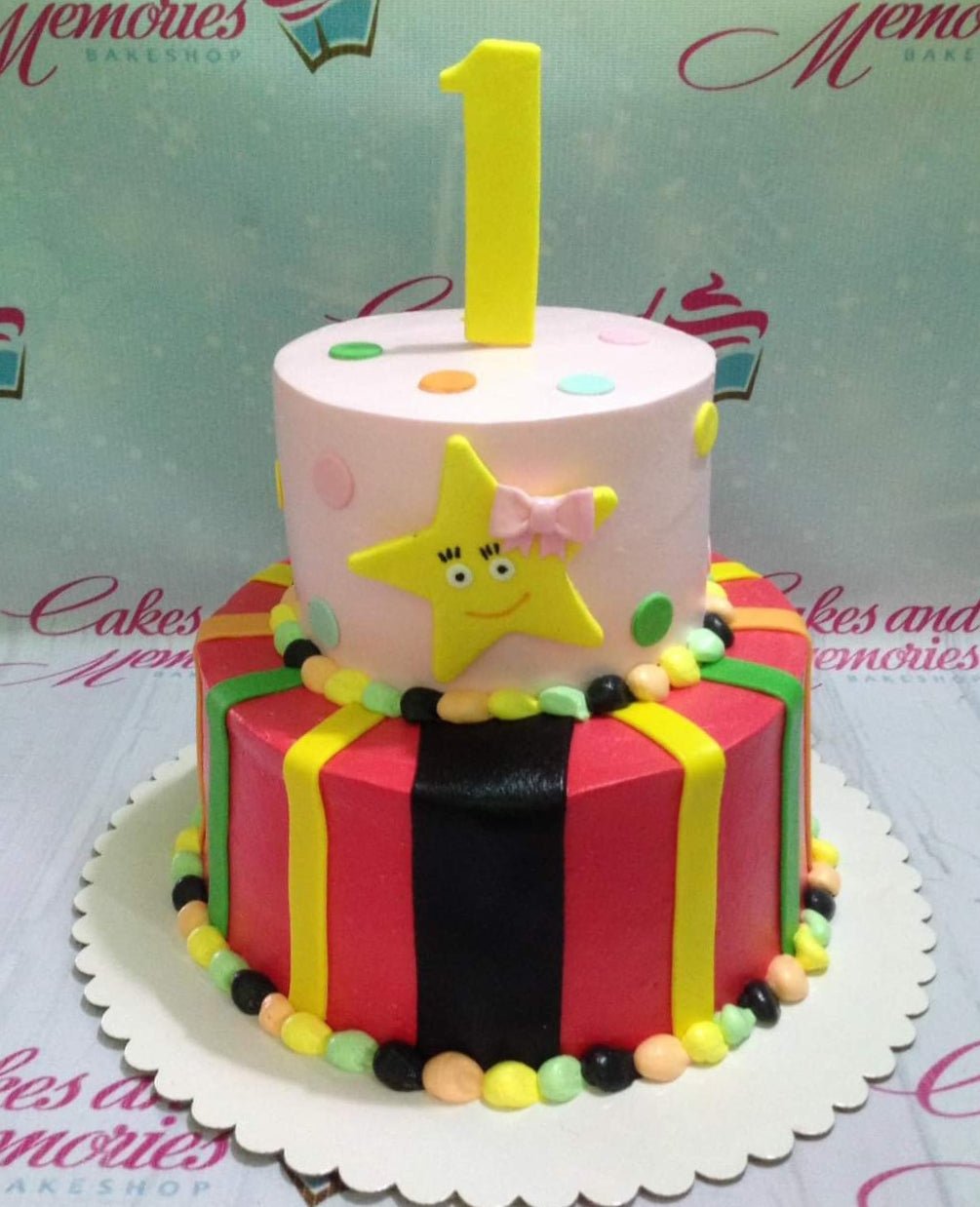 Two-tier birthday cake with a pink top tier featuring a smiling yellow star and a red bottom tier with colorful vertical stripes. Two-tier birthday cake with a pink top tier featuring a smiling yellow star and a red bottom tier with colorful vertical stripes.
