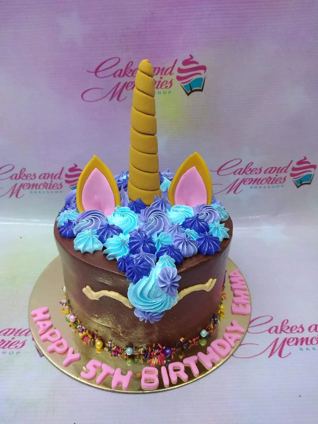 Chocolate unicorn cake with a gold horn, pink ears, and blue and purple icing rosettes on a gold board with a birthday message.