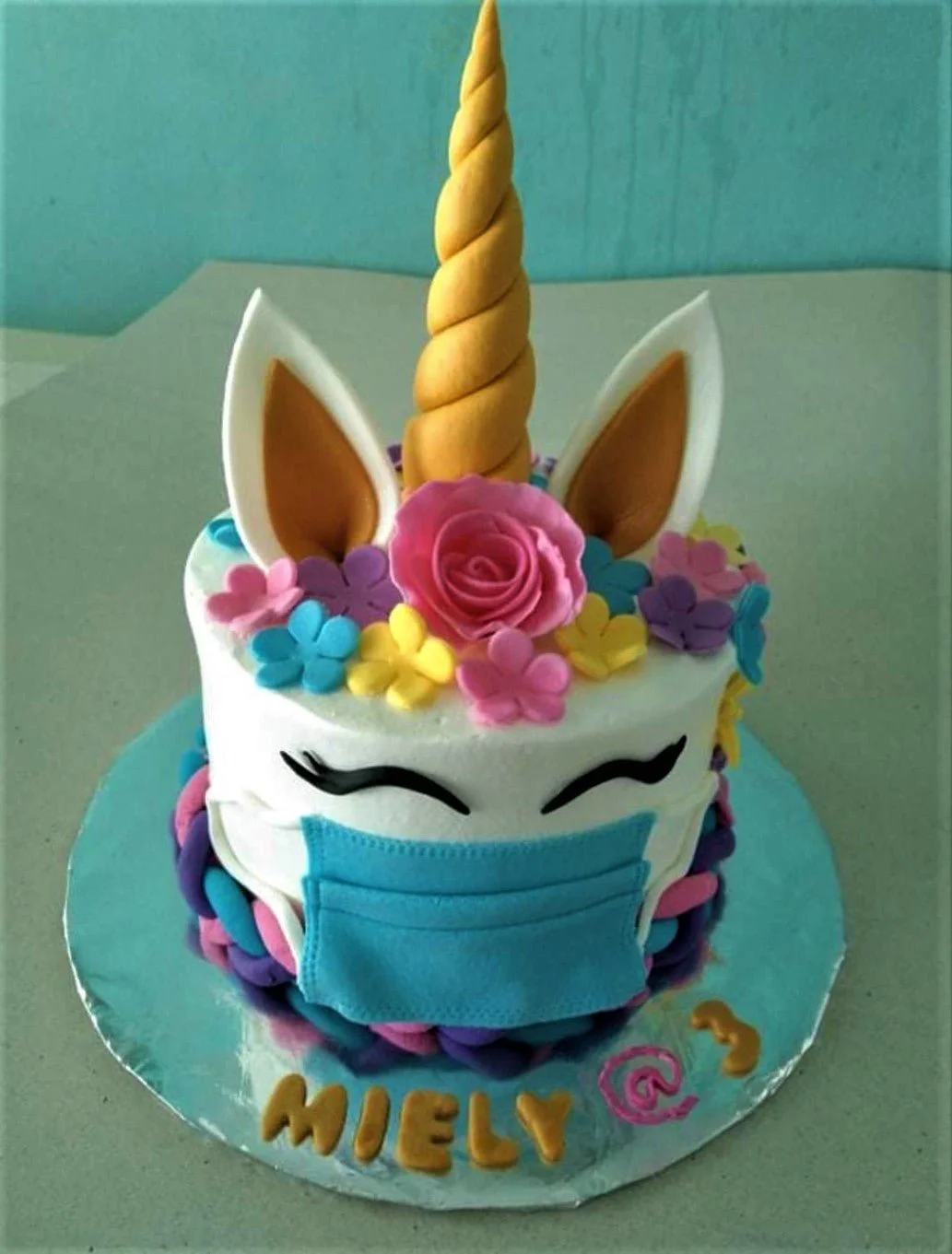 White unicorn cake wearing a blue face mask with a gold horn, pink rose, and colorful fondant flowers.