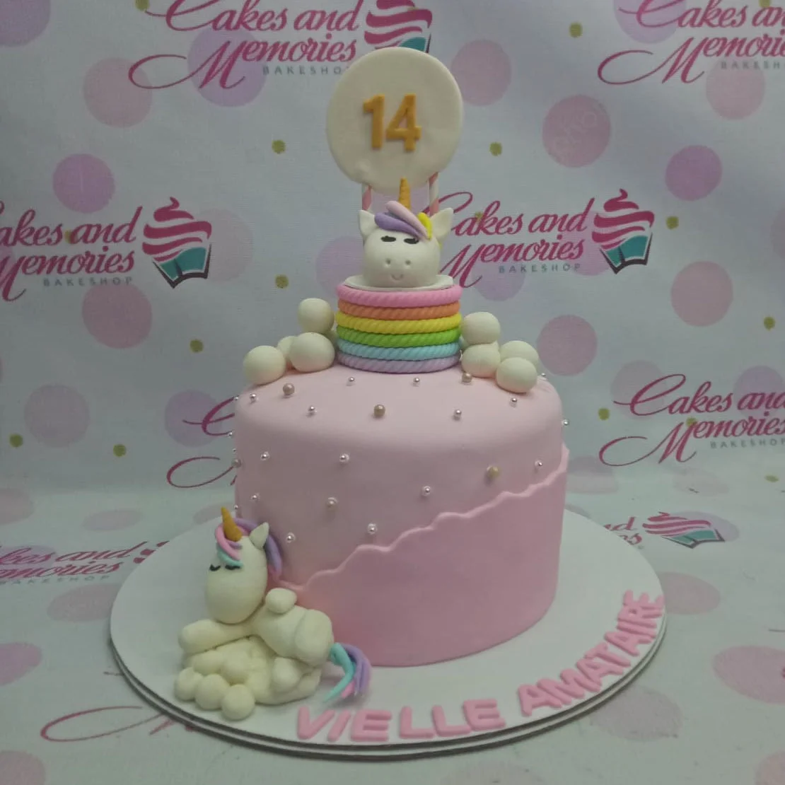 Pink fondant unicorn cake with rainbow rope border, white clouds, and a 14th birthday topper.