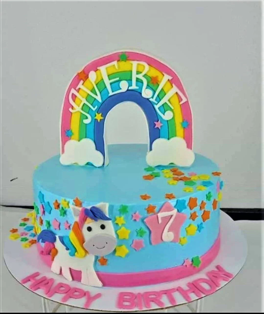 Unicorn Rainbow Sky Blue Birthday Cake A light blue 1-tier Unicorn Rainbow cake, featuring a rainbow, white unicorn, and pink number 7 topper, with colorful stars.