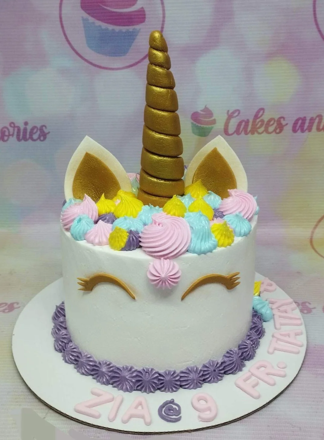 White unicorn cake with gold horn and ears, colorful icing rosettes, and purple star border on a white board.