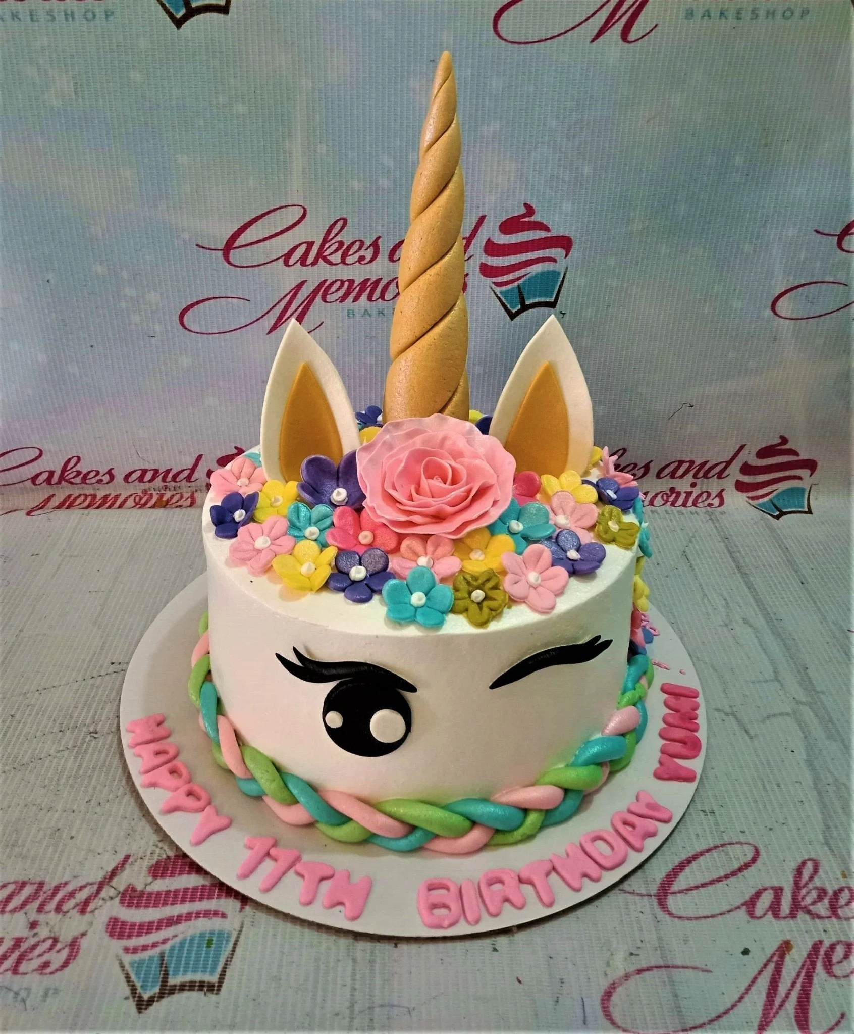White unicorn cake with a gold horn, pink rose, and colorful fondant flowers, featuring a braided pastel border and birthday message.