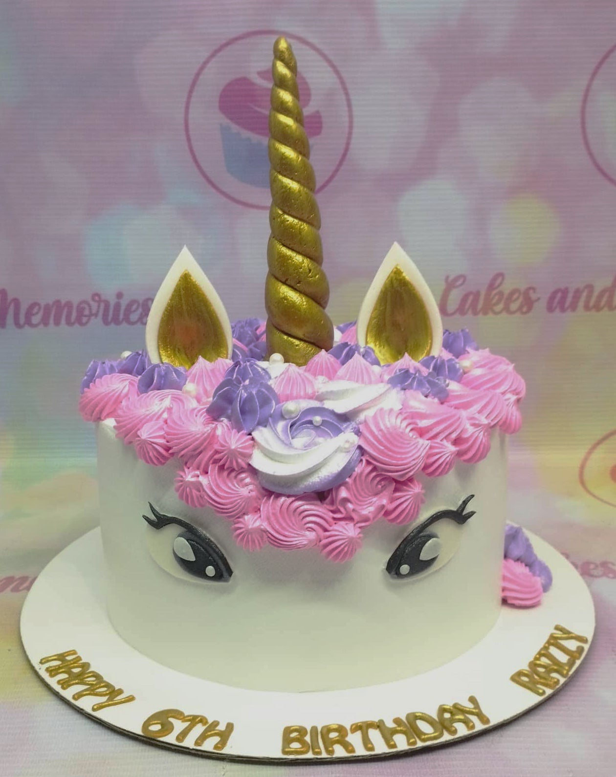 White unicorn birthday cake with pink and purple icing rosettes, gold horn and ears, and gold message on the base board. White unicorn birthday cake with pink and purple icing rosettes, gold horn and ears, and gold message on the base board.