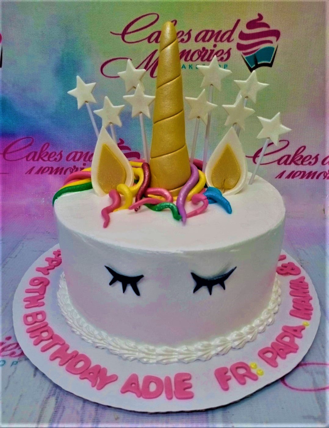 White unicorn cake with a gold horn, white ears, colorful fondant mane, and white star toppers on a pink message board. White unicorn cake with a gold horn, white ears, colorful fondant mane, and white star toppers on a pink message board.