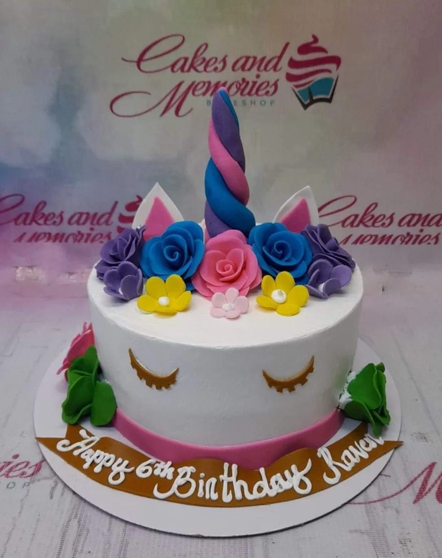 White unicorn cake with a colorful twisted horn, fondant ears, and a crown of vibrant pink, blue, purple, and yellow edible flowers.