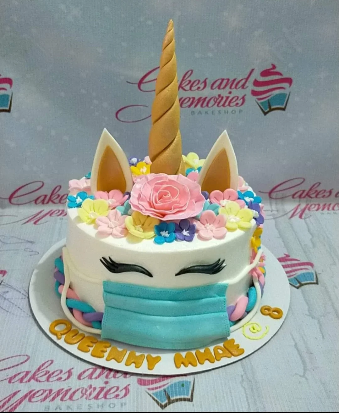 White unicorn cake with a gold horn, pink rose, colorful flowers, and a blue face mask decoration.