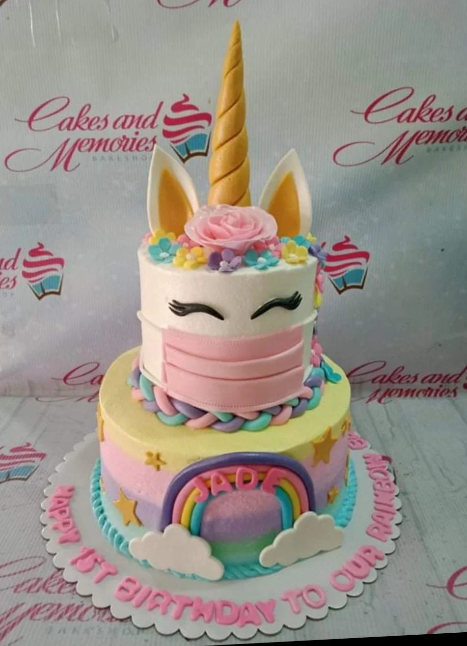 Two-tier unicorn birthday cake with a pink face mask, rainbow, and gold horn on a pastel gradient base.