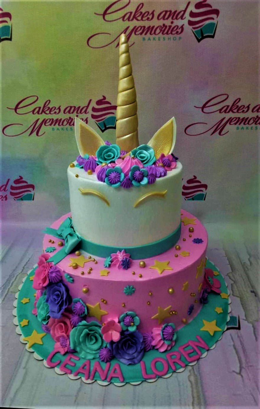 Two-tier unicorn themed cake with white and pink icing, decorated with colorful fondant flowers, gold stars, and a gold unicorn horn topper. Two-tier unicorn themed cake with white and pink icing, decorated with colorful fondant flowers, gold stars, and a gold unicorn horn topper.
