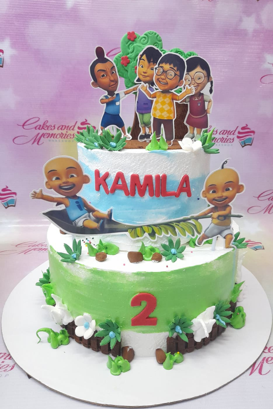 Two-tier Upin and Ipin themed birthday cake with green and blue icing, character printouts, and a red number 2. Two-tier Upin and Ipin themed birthday cake with green and blue icing, character printouts, and a red number 2.