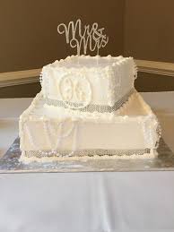 A white 2-tier cake featuring a 'Mr & Mrs' topper, piped icing swags, a piped monogram, and a silver dragee border. A white 2-tier cake featuring a 'Mr & Mrs' topper, piped icing swags, a piped monogram, and a silver dragee border.