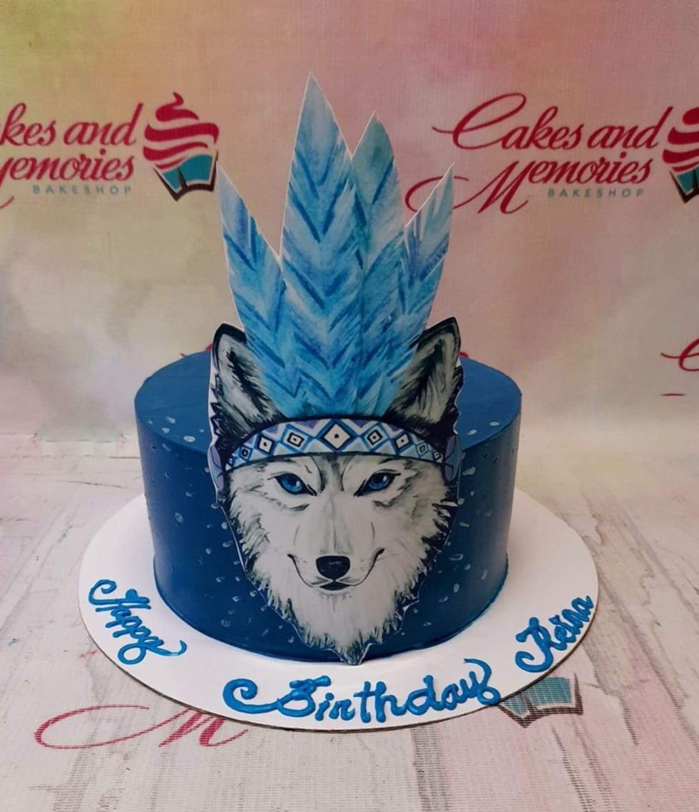 A deep blue single-tier cake featuring a large wolf printout topper with a blue feathered headdress and a birthday message on the base board. A deep blue single-tier cake featuring a large wolf printout topper with a blue feathered headdress and a birthday message on the base board.