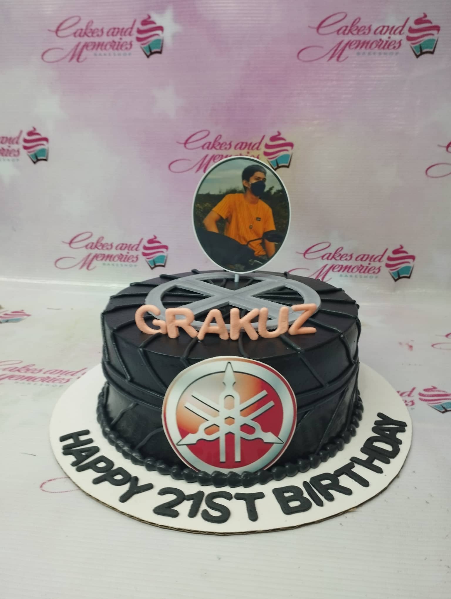 Black tire-themed birthday cake with a Yamaha logo and a photo printout topper for a 21st birthday celebration. Black tire-themed birthday cake with a Yamaha logo and a photo printout topper for a 21st birthday celebration.
