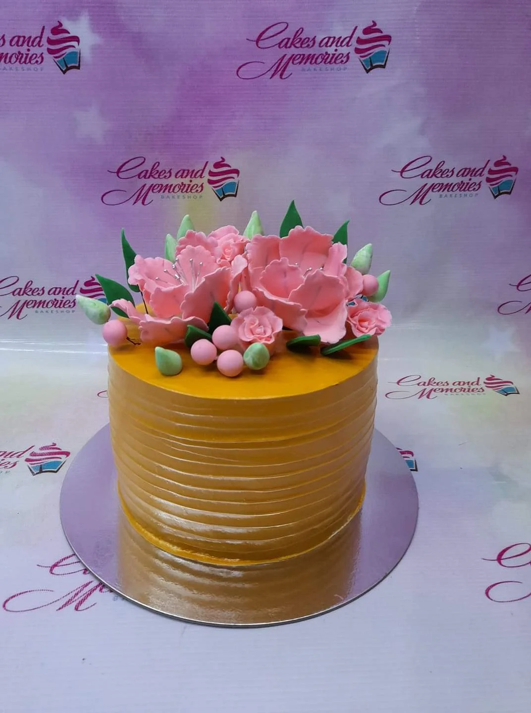 Yellow Peony Orange Floral Cake A yellow 1-tier cake with horizontal textured icing, featuring large pink gumpaste peonies, green leaves, small pink roses, and pink berries.