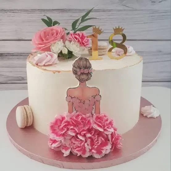 Elegant white 18th birthday cake featuring a printout of a girl in a pink floral dress with gold number toppers and fresh flowers.
