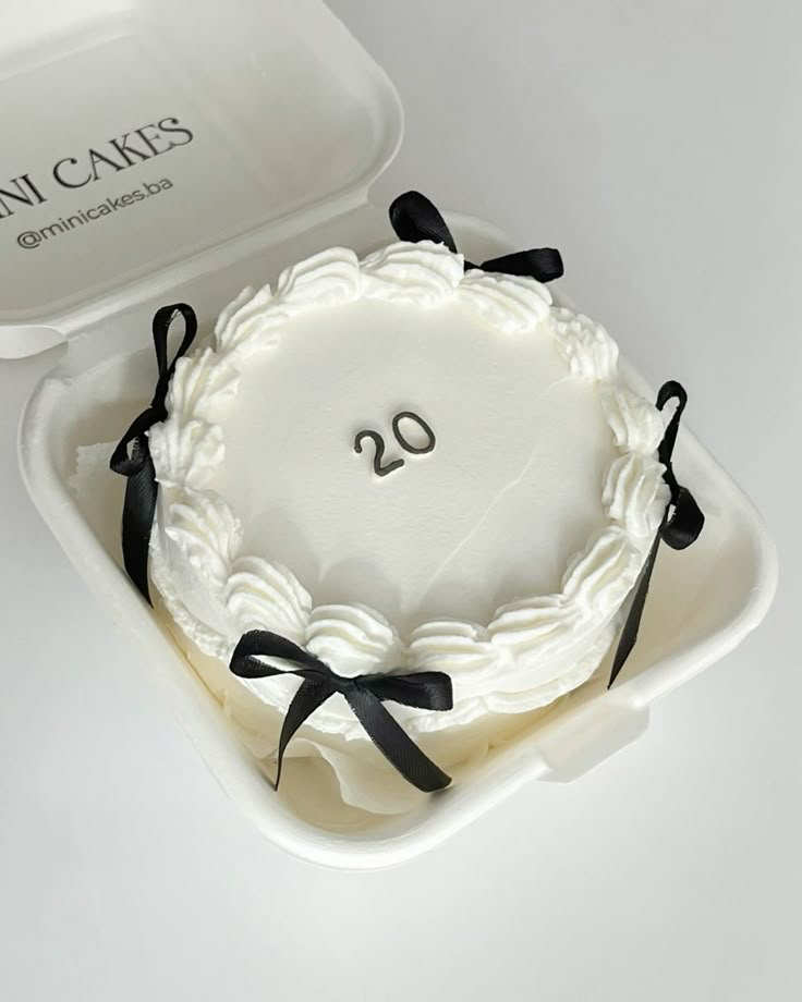 White minimalist bento cake with piped icing borders, black satin ribbons, and a silver number 20 topper in a white clamshell box.