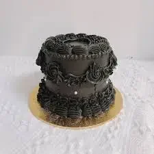 Elegant all-black vintage style cake with intricate piped icing ruffles, rosettes, and small white pearl accents on a gold base. Elegant all-black vintage style cake with intricate piped icing ruffles, rosettes, and small white pearl accents on a gold base.