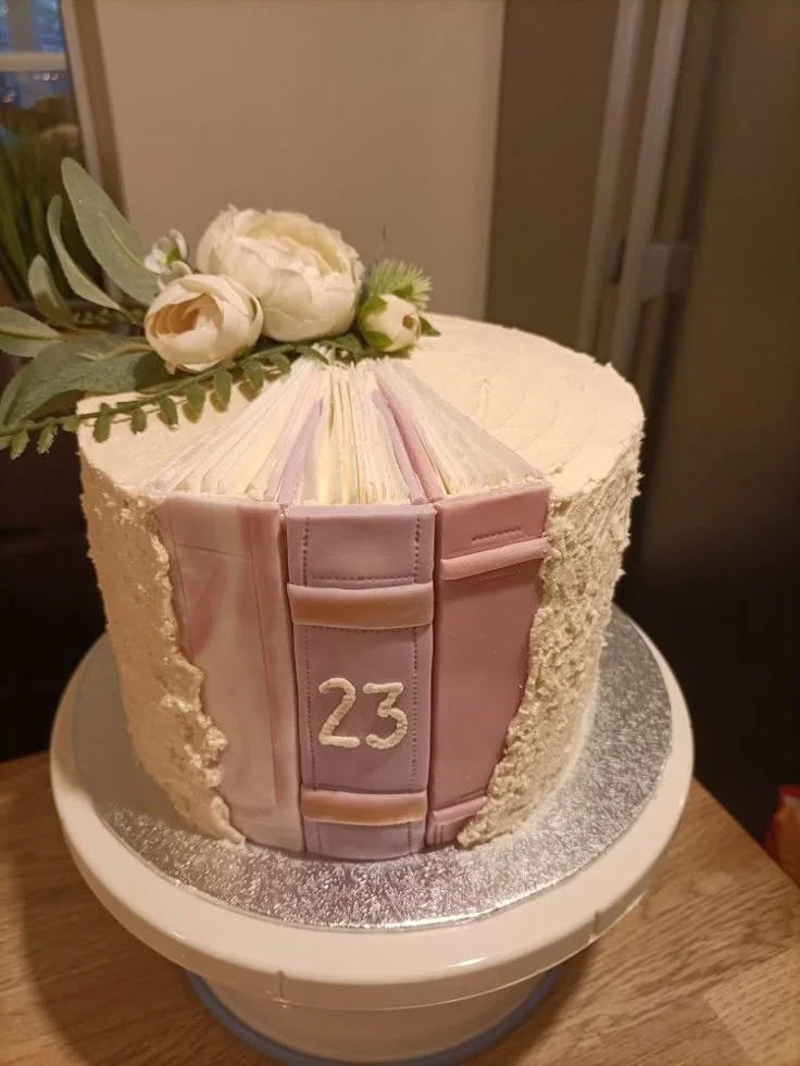 A cream-colored 1-tier cake designed to look like a stack of three books with white flowers and green leaves on top. A cream-colored 1-tier cake designed to look like a stack of three books with white flowers and green leaves on top.