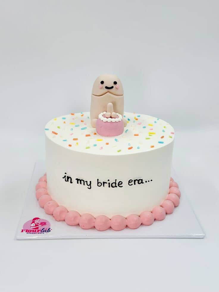 White minimalist cake with a humorous 3D fondant figure holding a small pink cake, decorated with colorful sprinkles and pink icing borders.