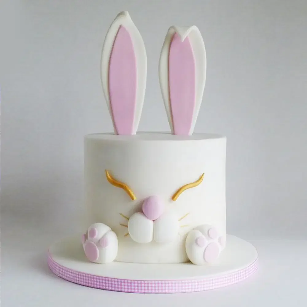 White fondant bunny cake with large pink and white ears, golden closed eyes, and 3D paws on a pink checkered base. White fondant bunny cake with large pink and white ears, golden closed eyes, and 3D paws on a pink checkered base.