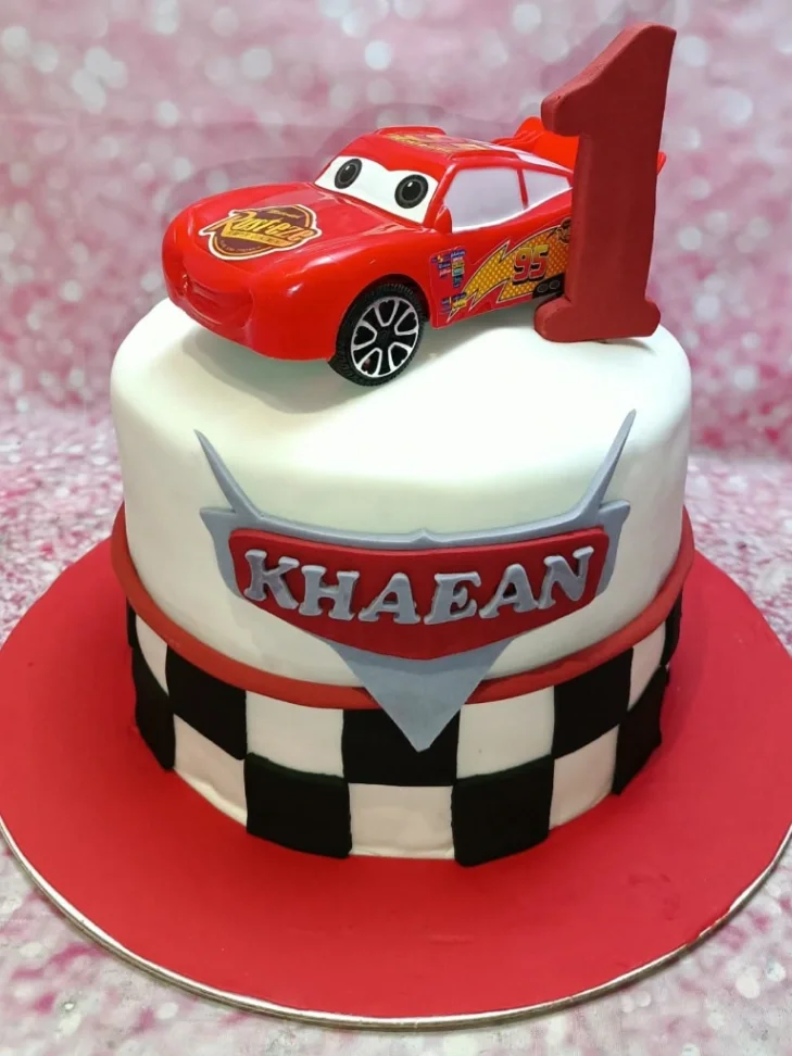 Cars Lightning McQueen-Inspired Birthday Cake A white fondant Cars themed cake with a red Lightning McQueen toy car, a red number one, and a checkered racing flag pattern.