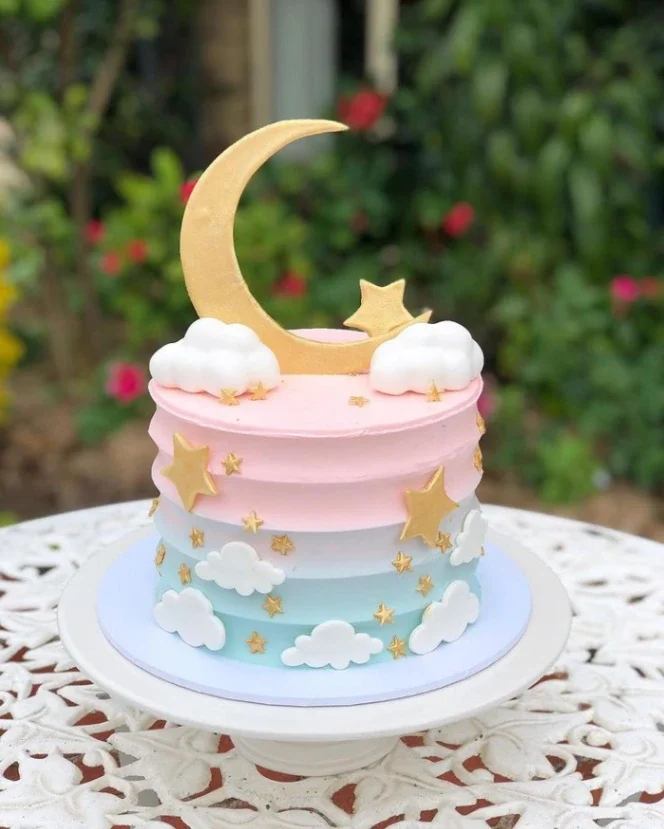 A pink and light blue gradient cake with white fondant clouds, gold stars, and a large gold crescent moon topper.
