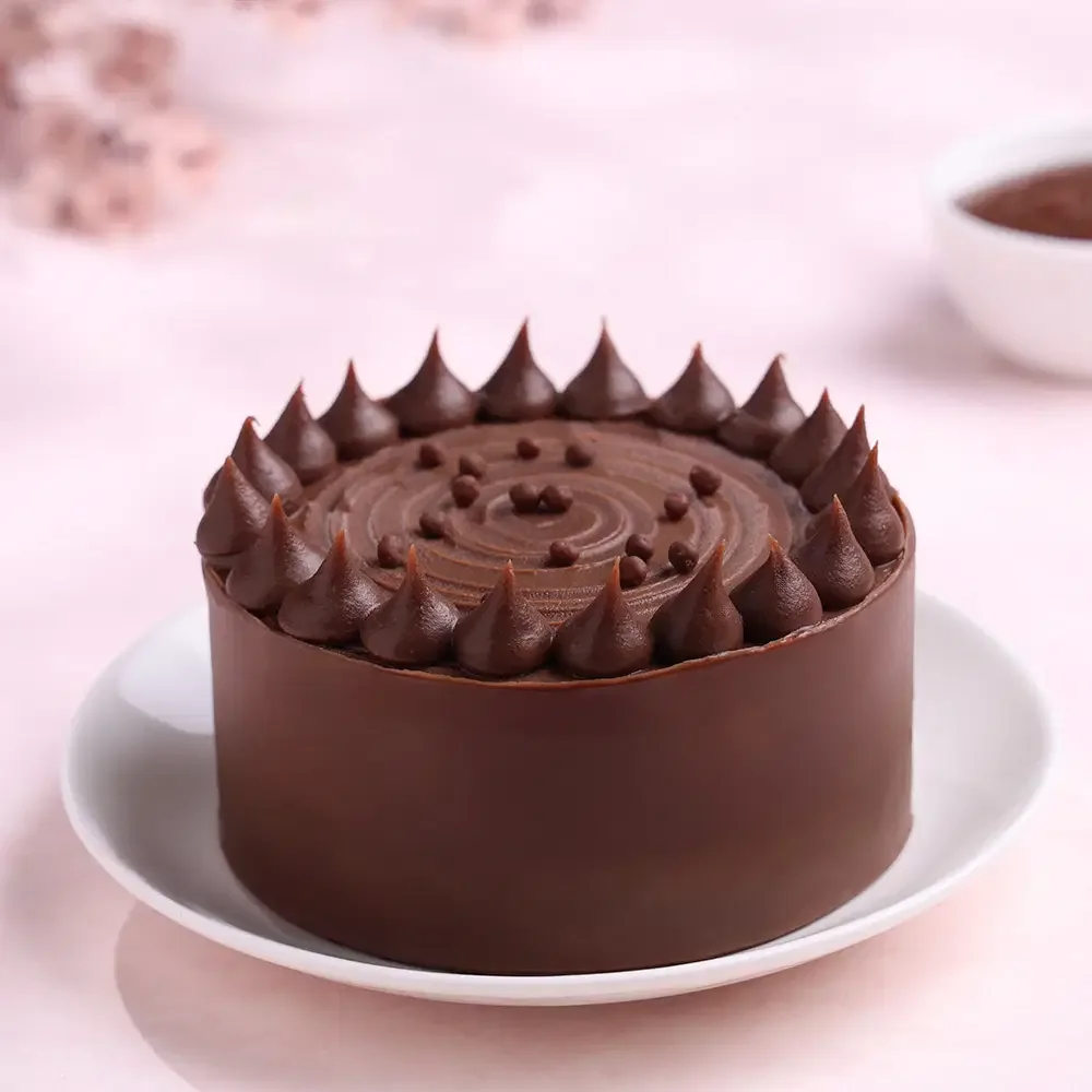 A smooth chocolate cake with dark chocolate icing dollops and small chocolate pearls on top, presented on a white plate. A smooth chocolate cake with dark chocolate icing dollops and small chocolate pearls on top, presented on a white plate.
