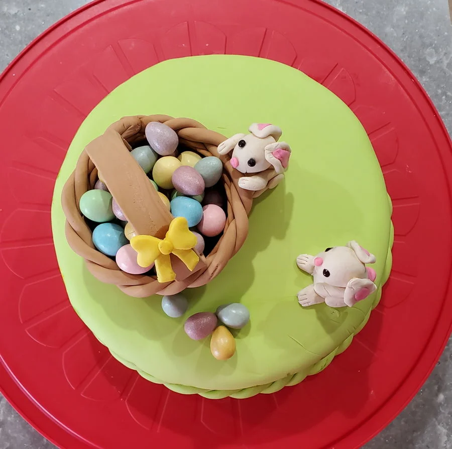 A light green fondant cake featuring a brown gumpaste basket filled with colorful edible eggs and two small white fondant bunnies. A light green fondant cake featuring a brown gumpaste basket filled with colorful edible eggs and two small white fondant bunnies.