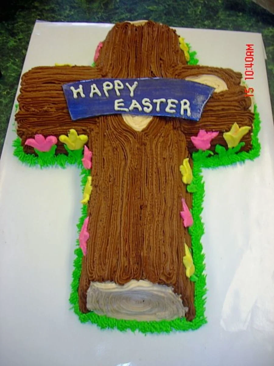 A brown cross-shaped cake with wood-like icing texture, pink and yellow flowers, and a blue banner reading Happy Easter. A brown cross-shaped cake with wood-like icing texture, pink and yellow flowers, and a blue banner reading Happy Easter.