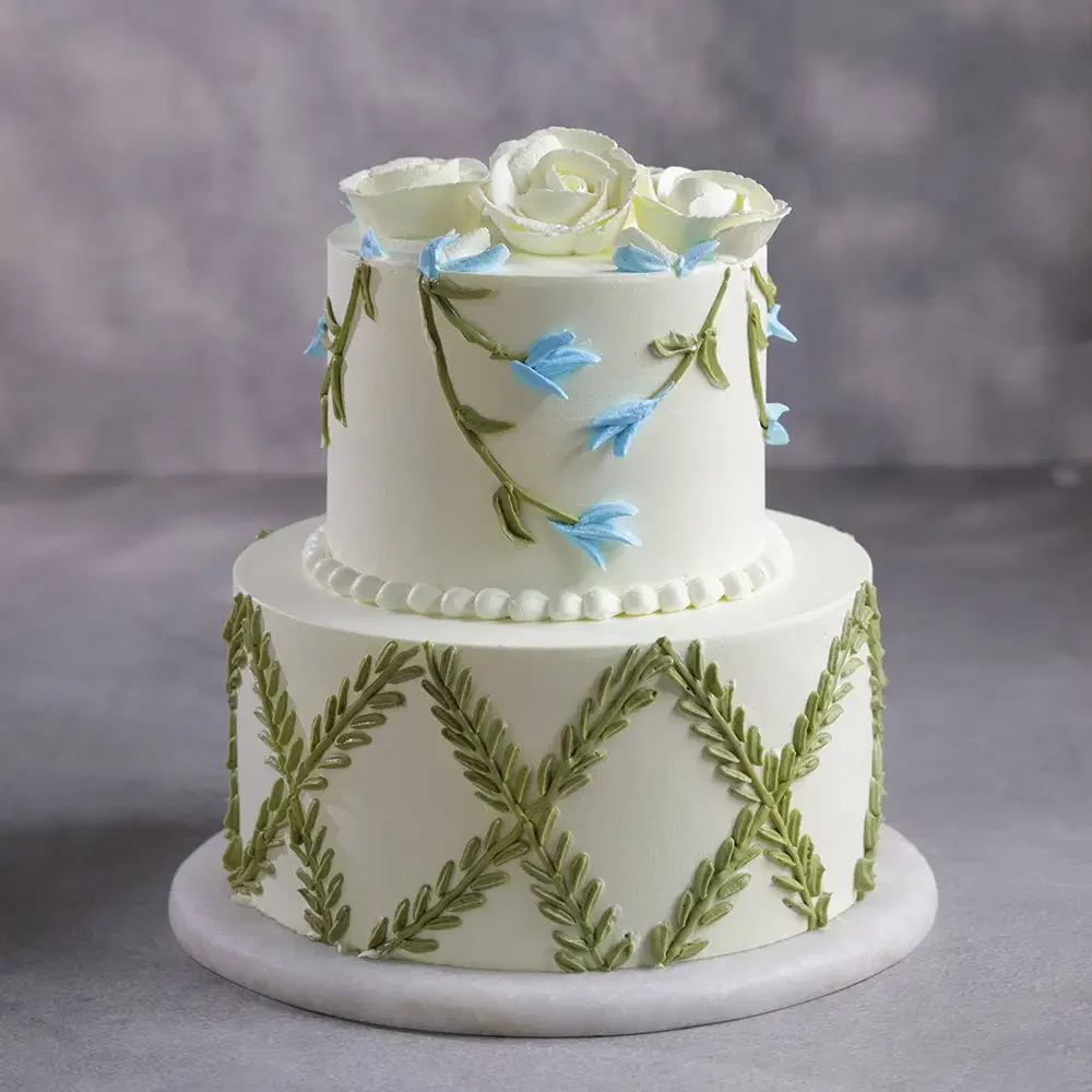 Floral Lattice White 2-Tier Cake White 2-tier soft icing cake with piped green leaf lattice, blue floral vines, and white rose toppers