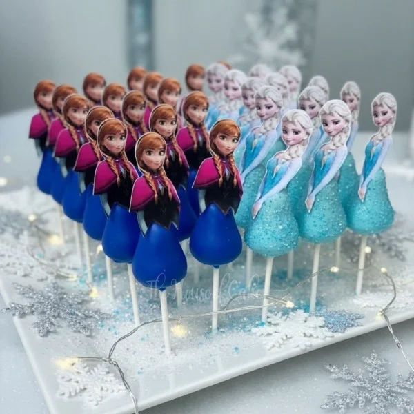 Frozen-Inspired Cake Pops Cupcakes Blue and white Frozen themed cake pops featuring printed Anna and Elsa character toppers on a tray with silver snowflake accents