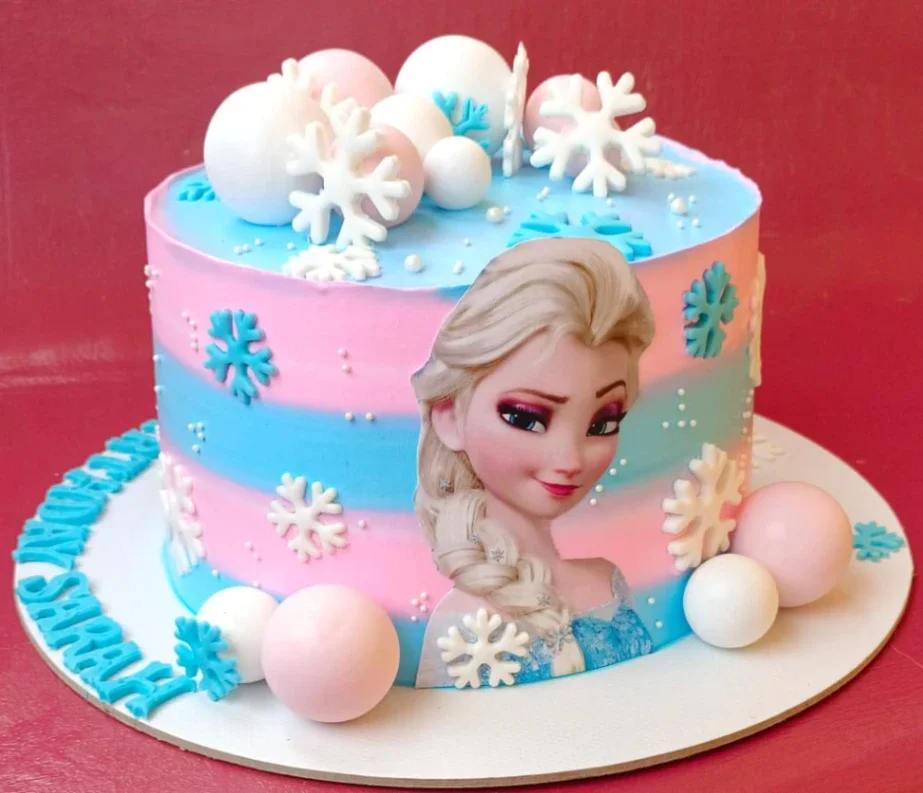 Frozen Elsa themed birthday cake with pink and blue gradient icing, white snowflakes, and pink and white fondant spheres.