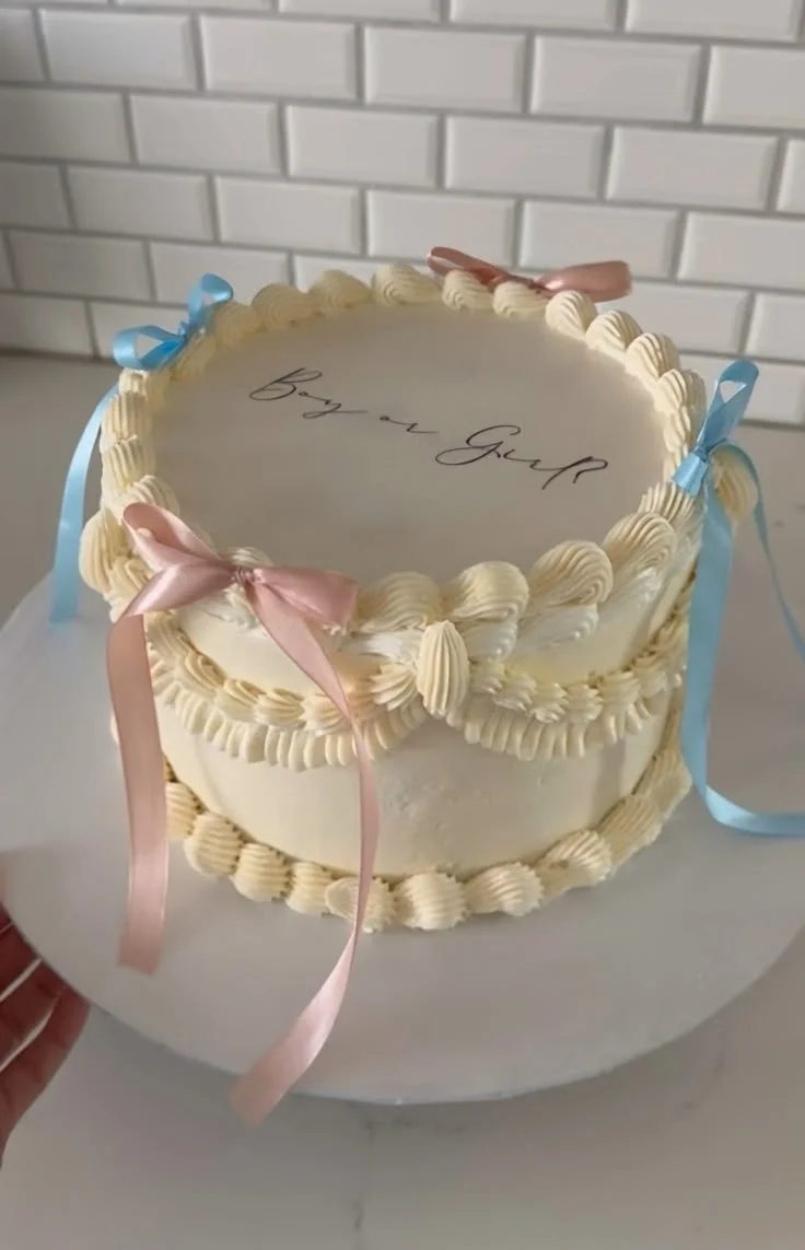 Elegant cream-colored vintage-style gender reveal cake with pink and blue satin ribbons and 'Boy or Girl' icing text.