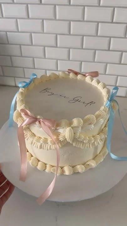 Elegant white vintage-style gender reveal cake with intricate cream piping, pink and blue satin ribbons, and 'Boy or Girl' text.