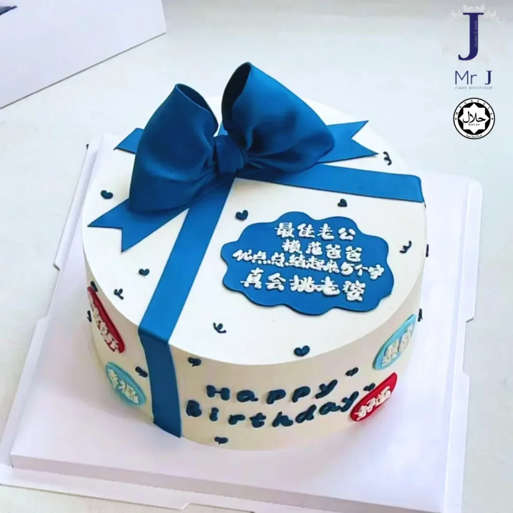 Gift Box White Bow Birthday Cake White 1-tier cake with smooth soft icing, a large blue fabric bow on top, blue ribbon strips, and colorful circular plaques on the sides