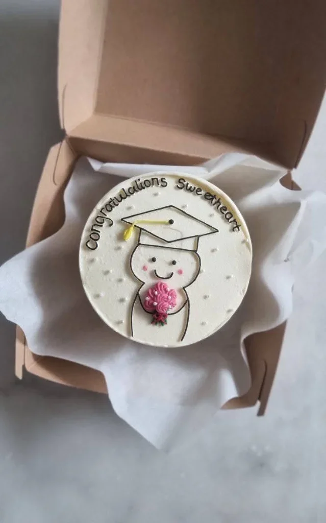 White bento cake with a hand-drawn minimalist graduation character holding a pink bouquet and a black graduation cap. White bento cake with a hand-drawn minimalist graduation character holding a pink bouquet and a black graduation cap.