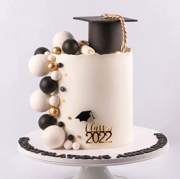 Elegant white graduation cake with a black fondant graduation cap, gold tassel, and black, white, and gold decorative spheres. Elegant white graduation cake with a black fondant graduation cap, gold tassel, and black, white, and gold decorative spheres.